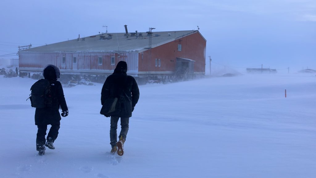 Two people walking through windy, snowy terrain towards a large, red building